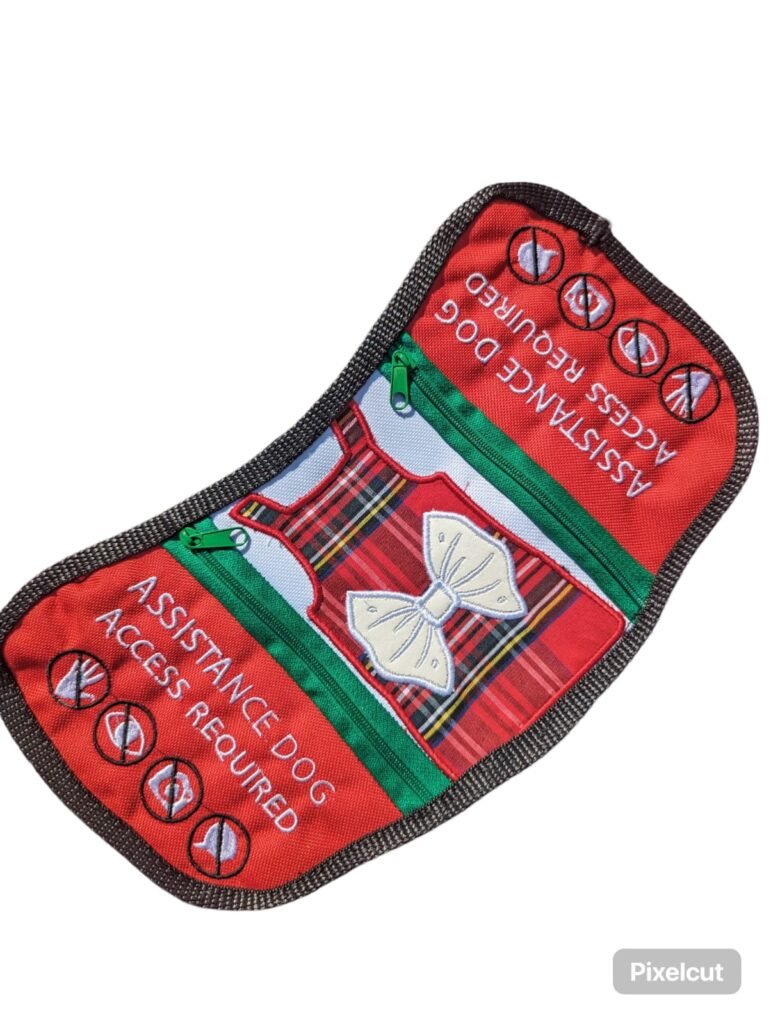 This vibrant cape, featuring red, black, and green with a tartan dress design, states "Assistance Dog, access required" with icons to ensure clear communication.